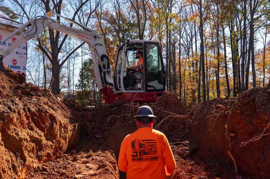 Two construction workers: one operates an excavator in a deep trench, while another views the trench from above.