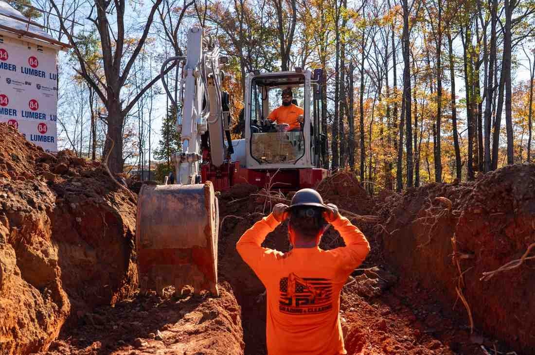 A construction worker in a safety vest stands in a dirt trench as another operates an excavator.