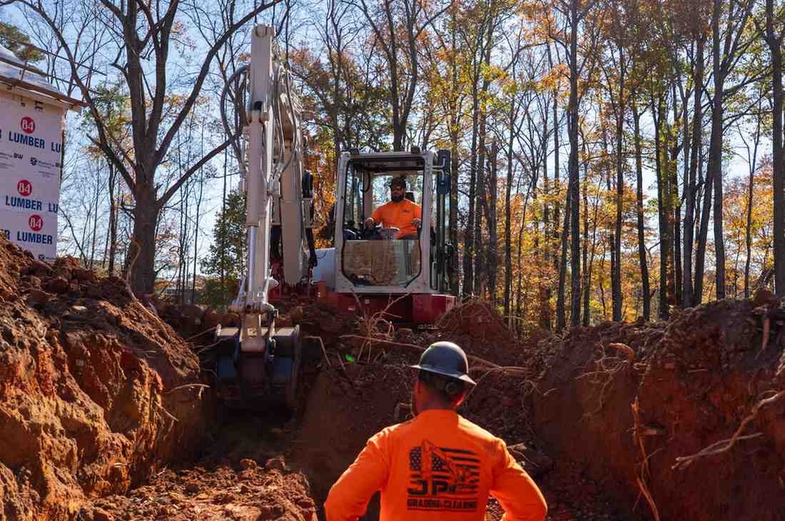 Two construction workers in orange shirts, one operating an excavator, and the other standing in a trench.