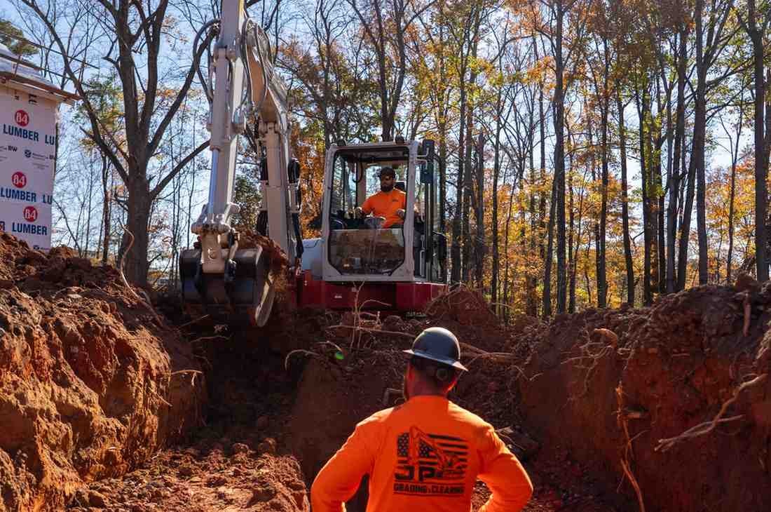 Workers on a construction site, one in a trench facing another operating an excavator, with a wooded area in the background.