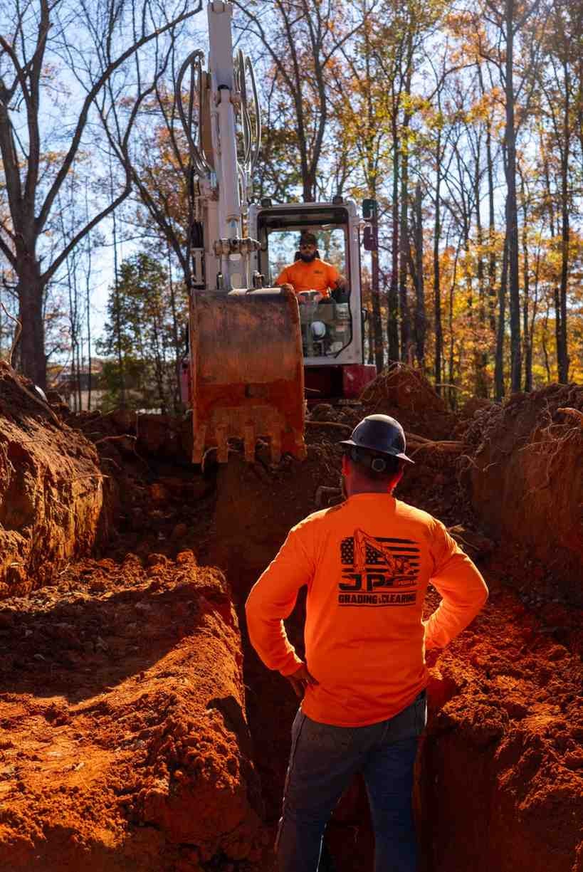 Two men work on a construction site; one operates an excavator, the other stands below in a trench.
