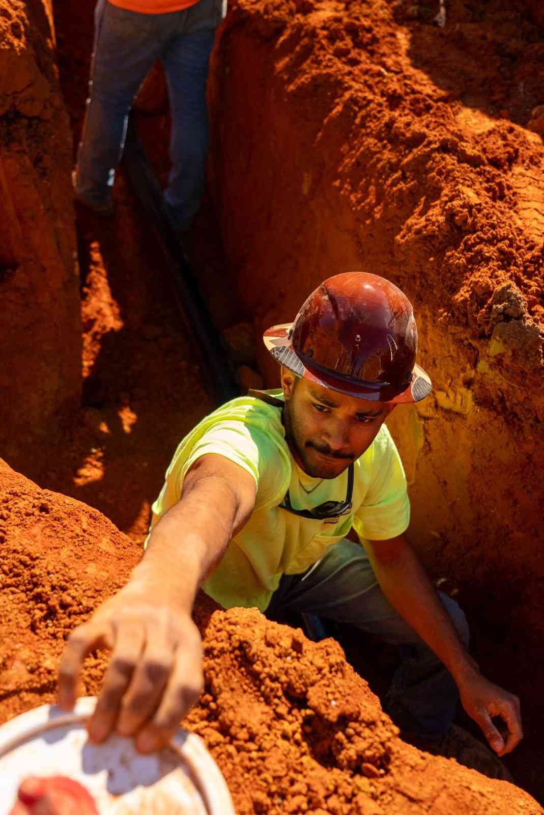 A man in a hard hat and yellow shirt crouches in a trench of red-orange dirt, reaching for a white plate.