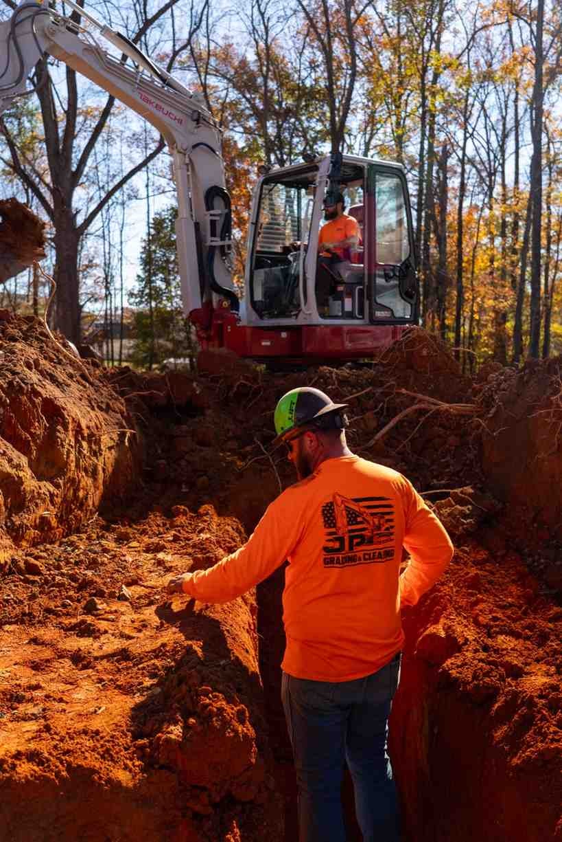 A construction worker in an orange shirt and green hard hat stands in a trench, looking at an excavator.