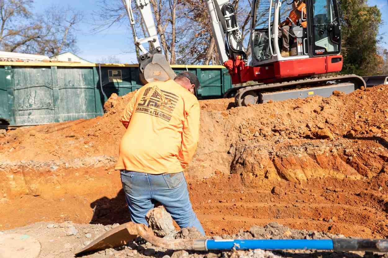 Construction worker in orange shirt and jeans digging a trench with an excavator and dumpster in the background.