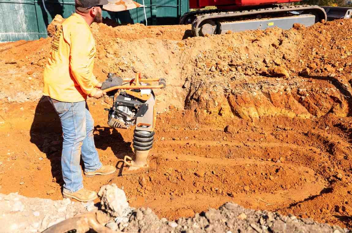 A person in an orange shirt and jeans operates a vibratory tamper in a trench of red-brown dirt.