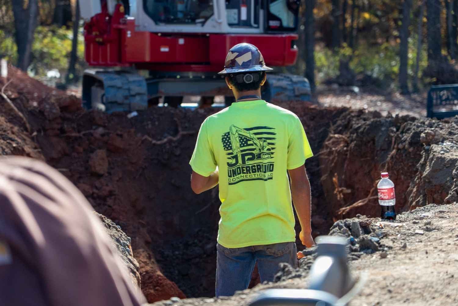 A construction worker in a yellow shirt and camouflage hard hat stands by a trench with excavation equipment.