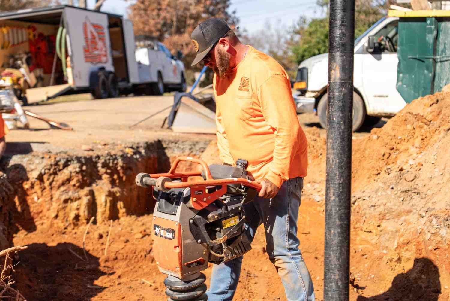 A construction worker in an orange shirt and jeans operates a tamping rammer in a dirt trench.