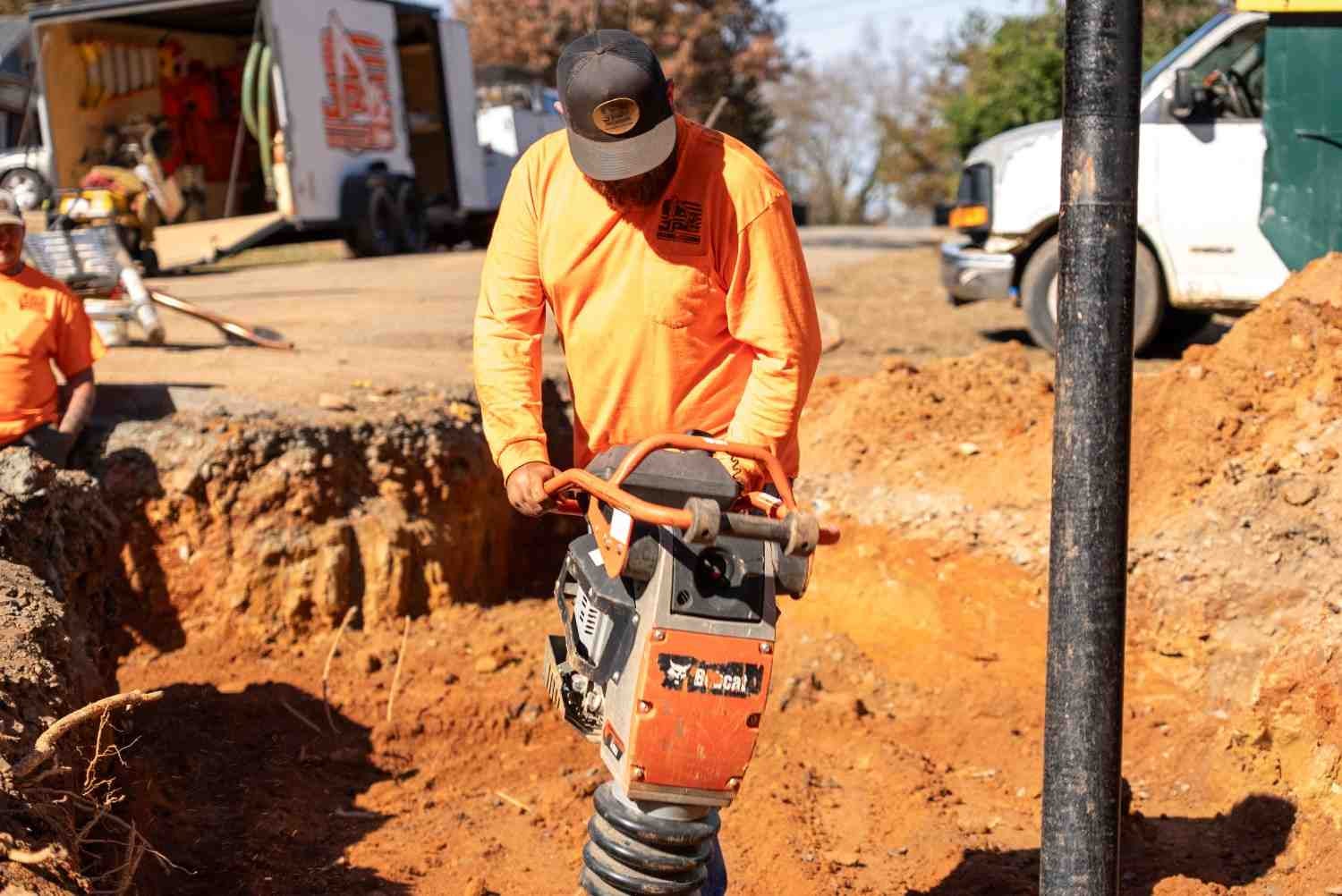 A construction worker in an orange shirt and hat operates a Bobcat tamper tool in a deep trench of red-brown dirt.