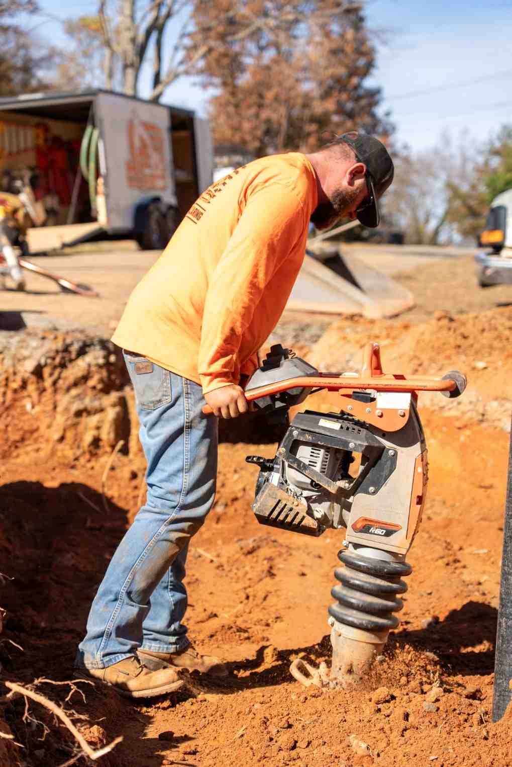 A construction worker in an orange shirt and jeans operates an orange and gray soil compactor in a trench.