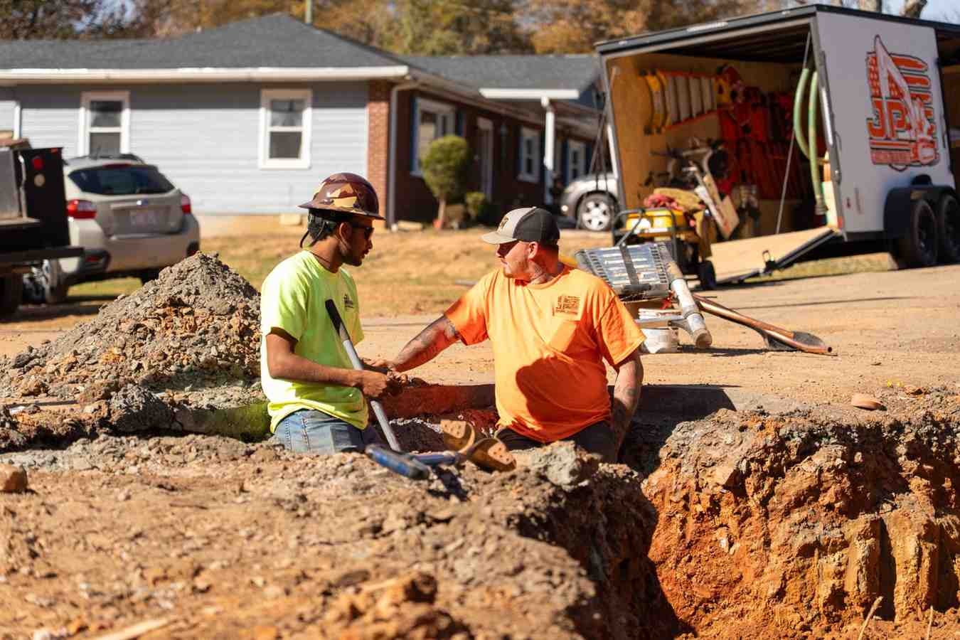 Two construction workers in a trench are talking. One wears a hardhat and holds a shovel, the other a cap.