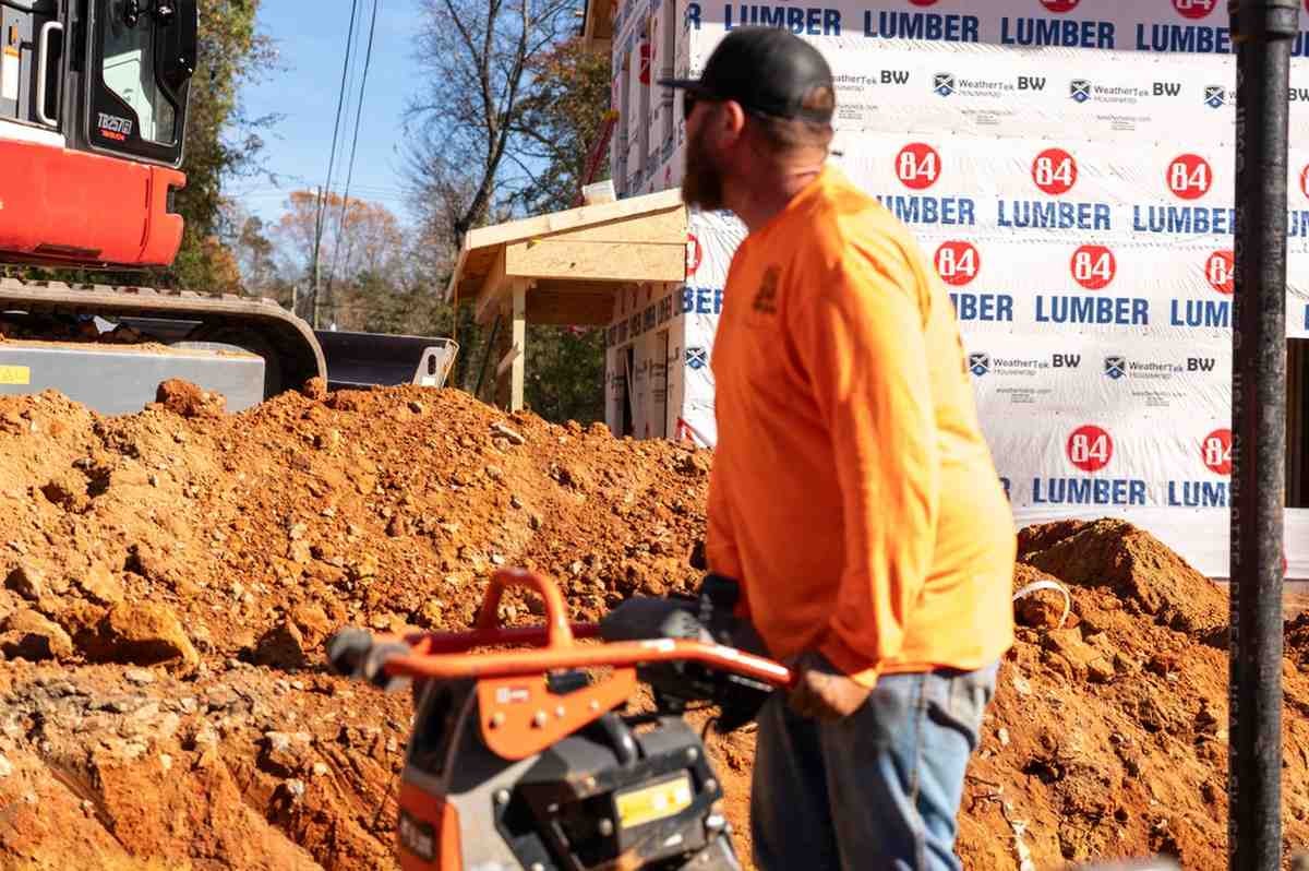 Construction worker in an orange shirt operating a compactor next to a pile of dirt, with a building under construction and an excavator in the background.