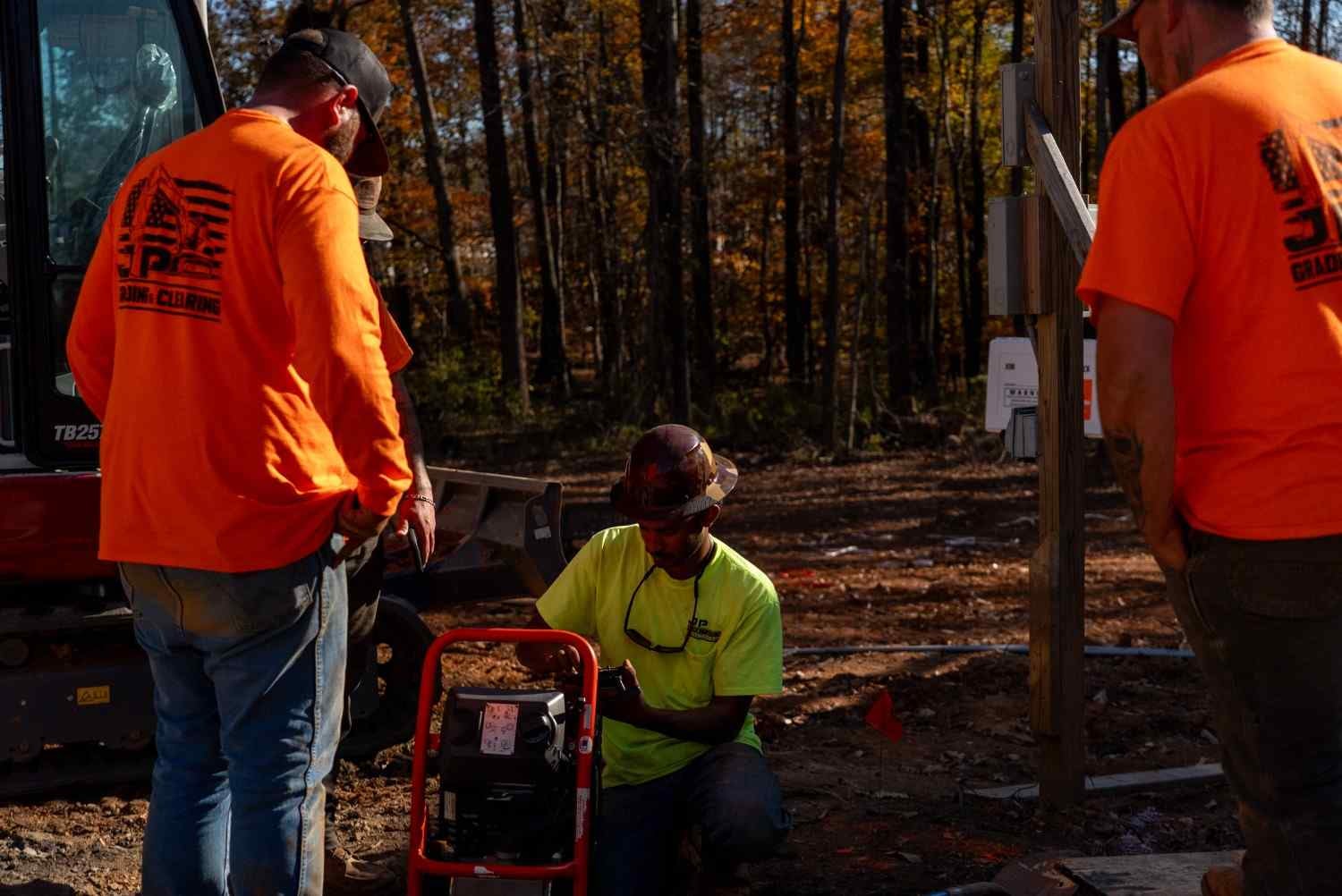 Three construction workers, two in orange and one in neon yellow, working on machinery at a wooded site.