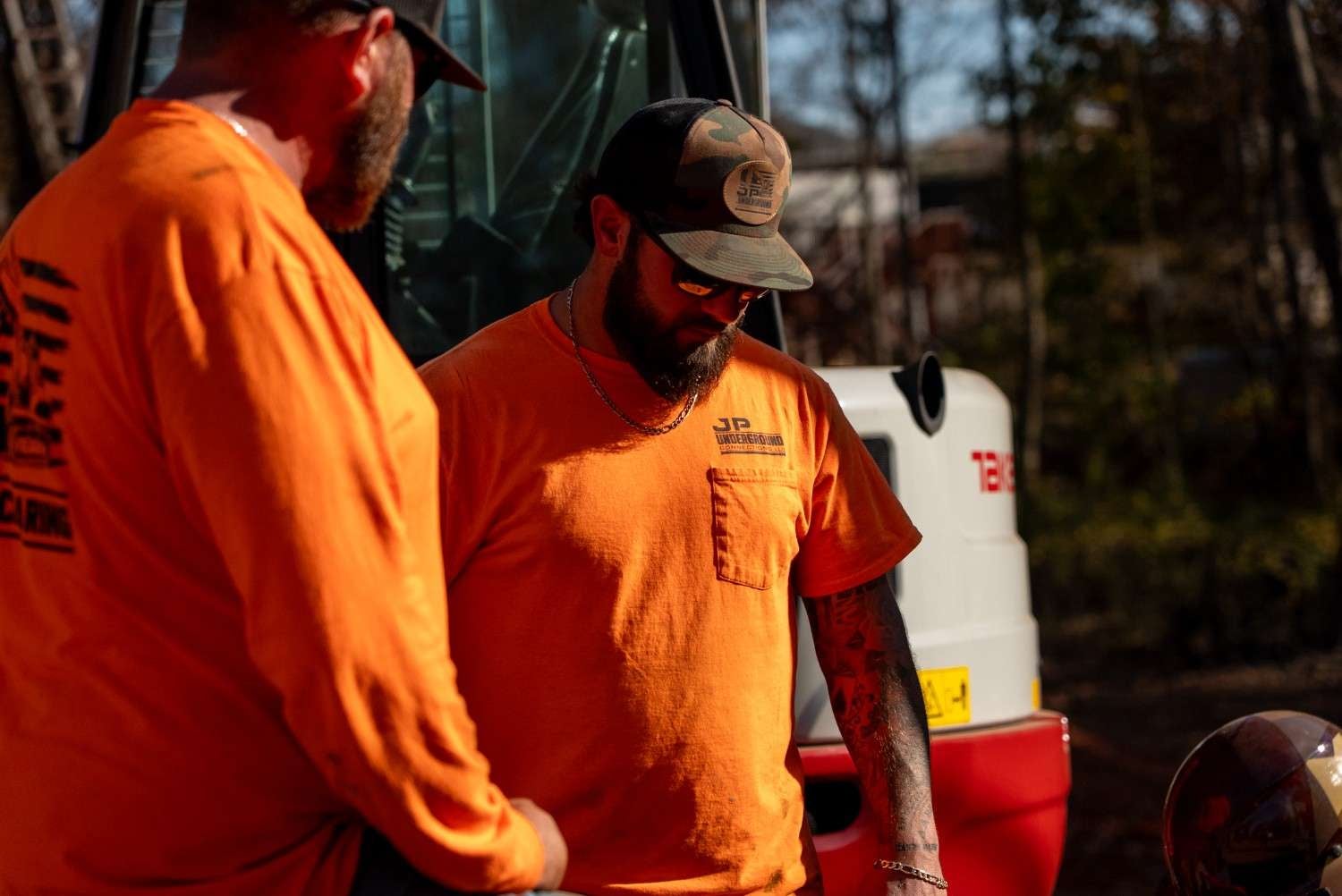 Two bearded men in orange shirts, one wearing sunglasses and a camo cap, outside with heavy machinery.