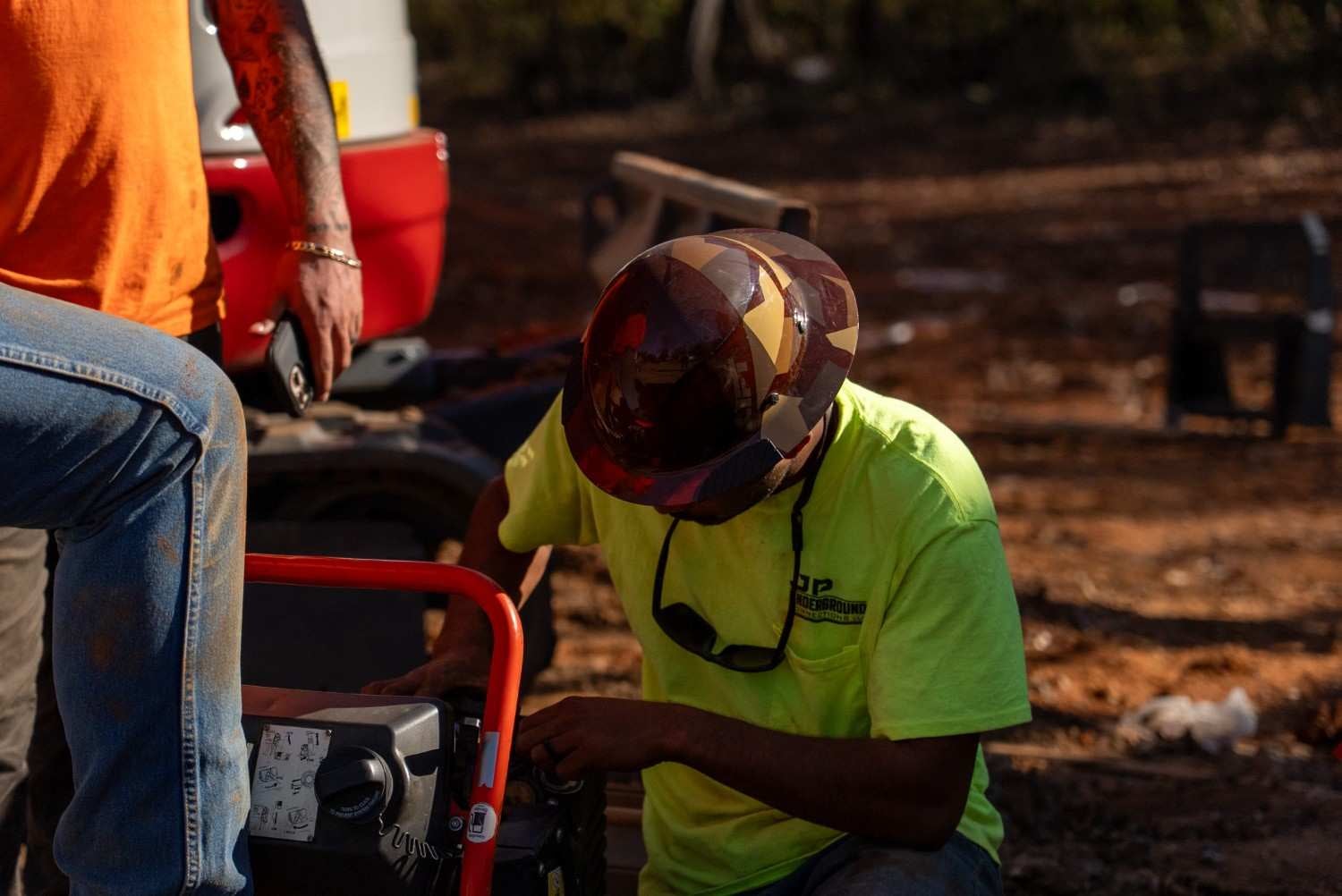 Construction worker in a patterned hard hat and safety green shirt crouches, adjusting equipment on a sunny day.