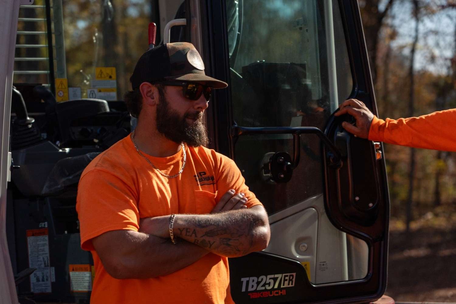 Man in orange work shirt, camo hat, and sunglasses with crossed arms next to heavy equipment.