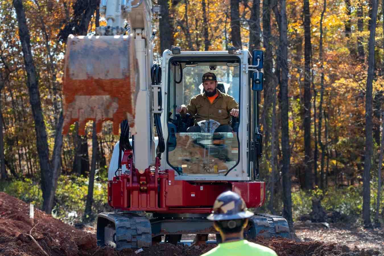 A man operates an excavator with a large, dirt-filled bucket, as another man in a hard hat watches.