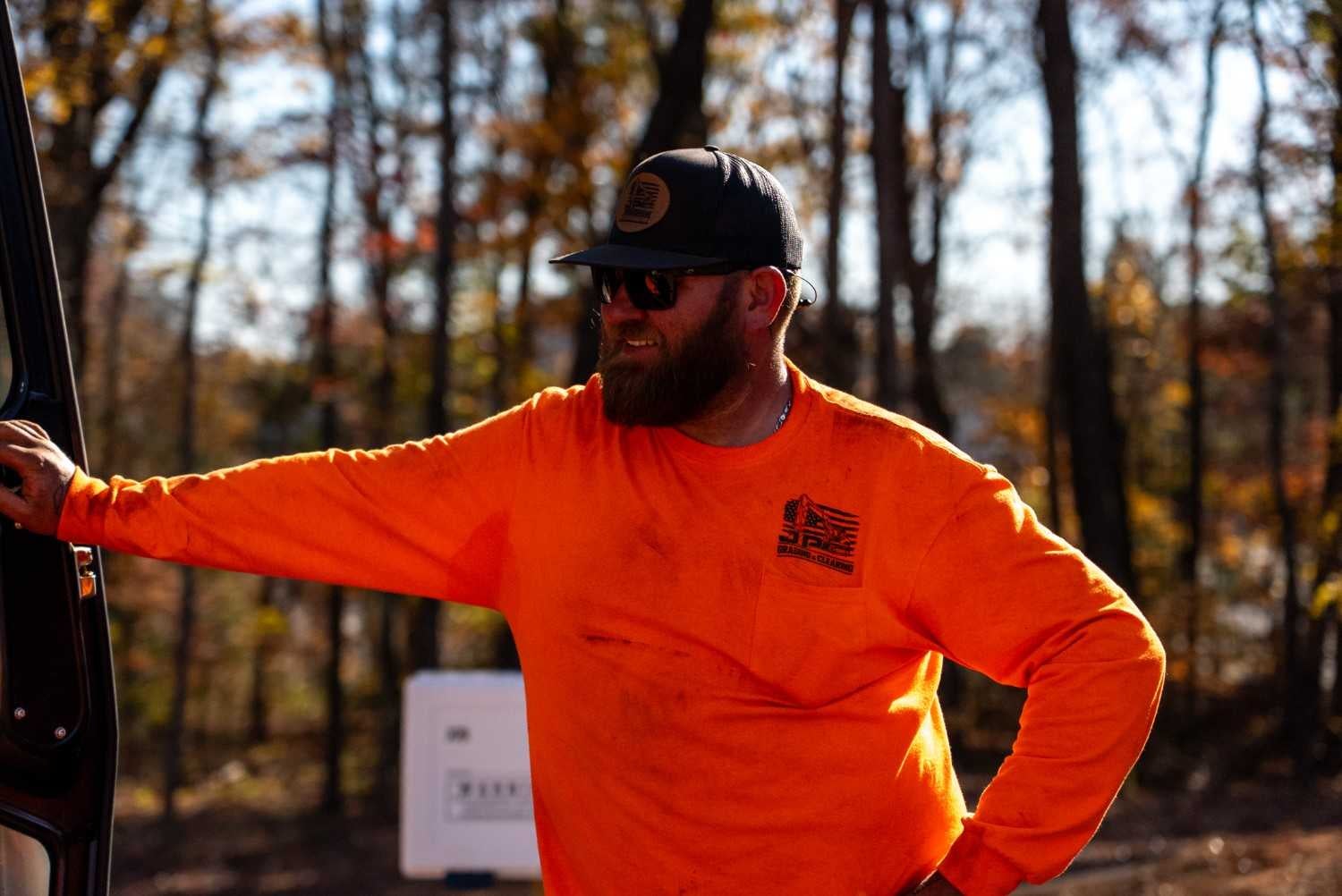 A man in an orange long-sleeved shirt, black cap, and sunglasses, leaning on a vehicle door in a sunlit forest.