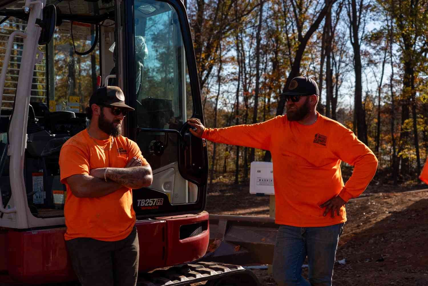Two construction workers in orange shirts and sunglasses stand by an excavator in a wooded area.