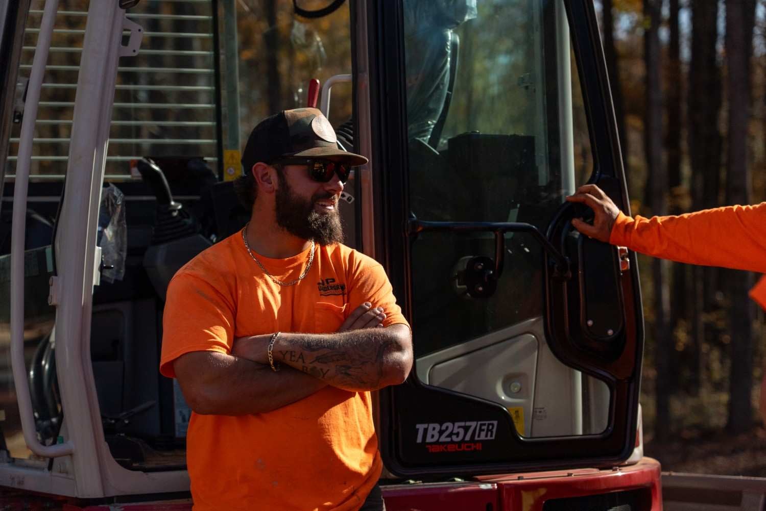 A bearded man in an orange shirt, sunglasses, and baseball cap stands with crossed arms next to heavy machinery.