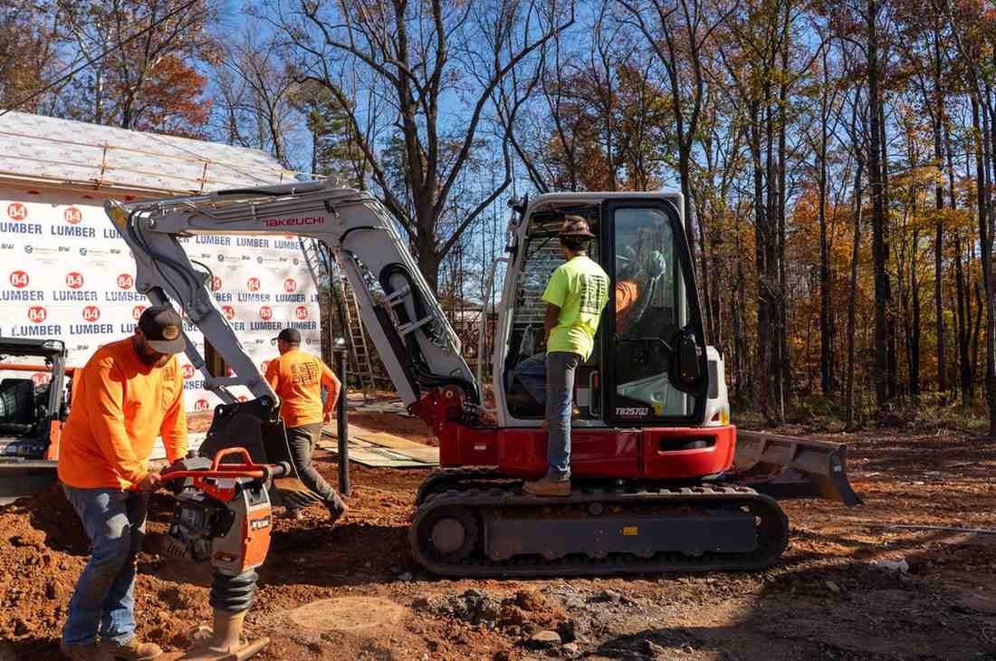 Construction workers on a building site, one operating a compactor, another an excavator, with a woodland background.