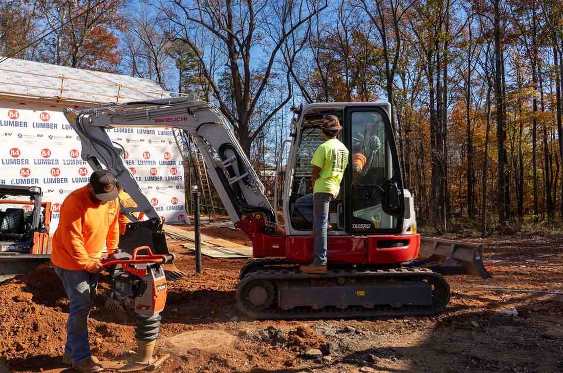 Two construction workers: one operates a jumping jack tamper, the other stands by an excavator at a worksite.
