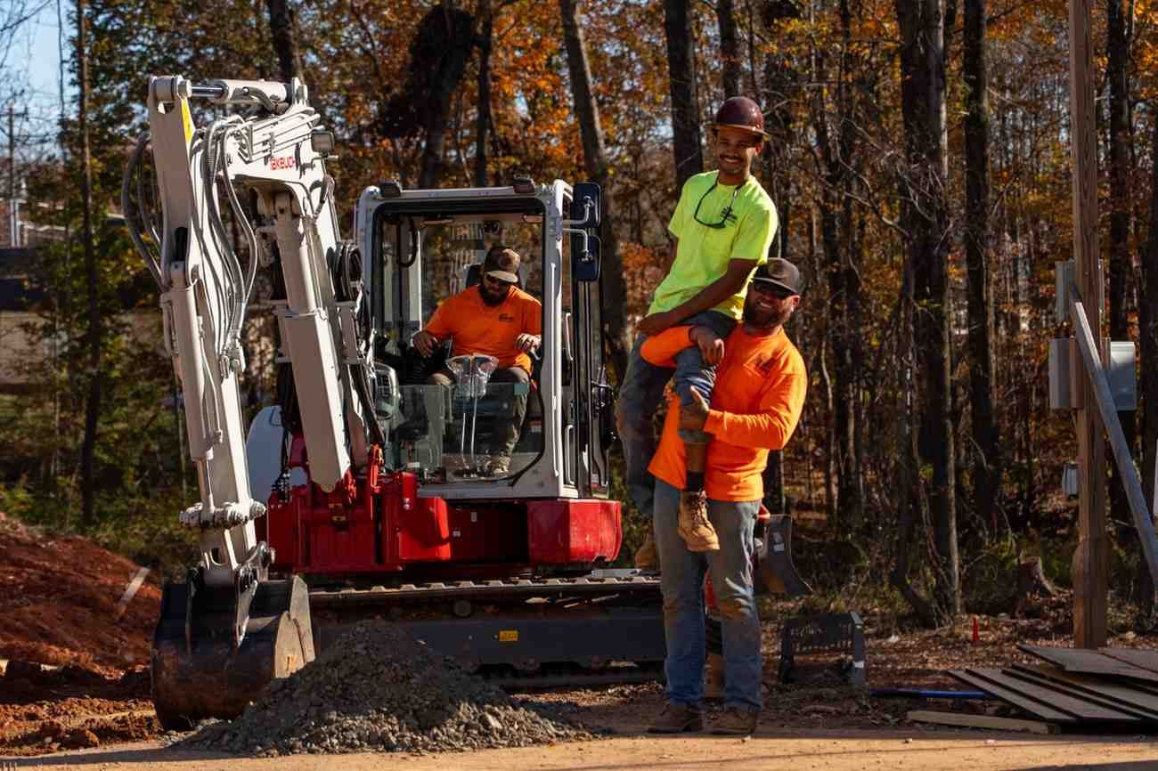Three construction workers, two smiling, pose with an excavator at a worksite with autumn trees in the background.