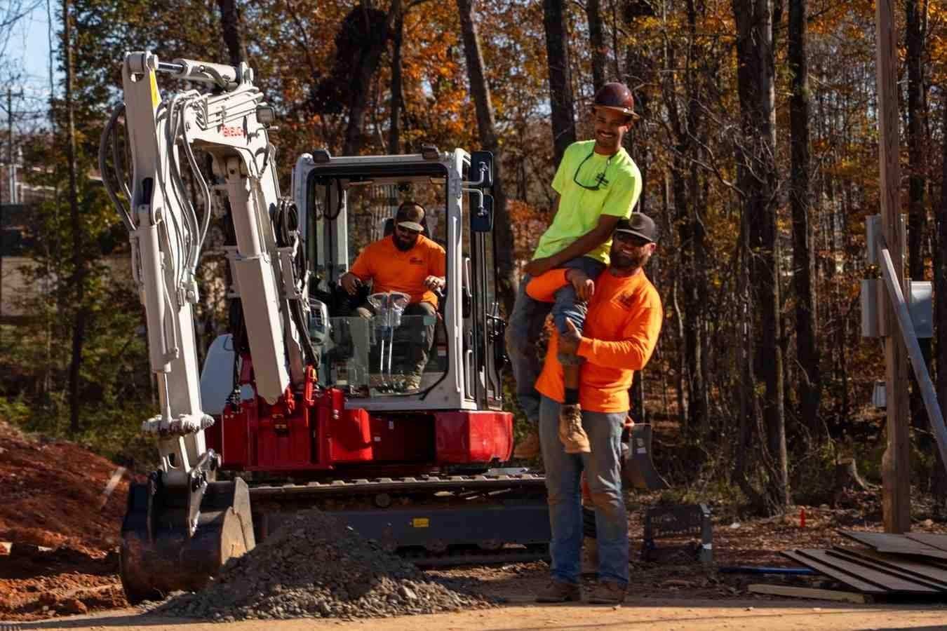 Three construction workers on a sunny day: one in an excavator, one carrying another on his shoulders.