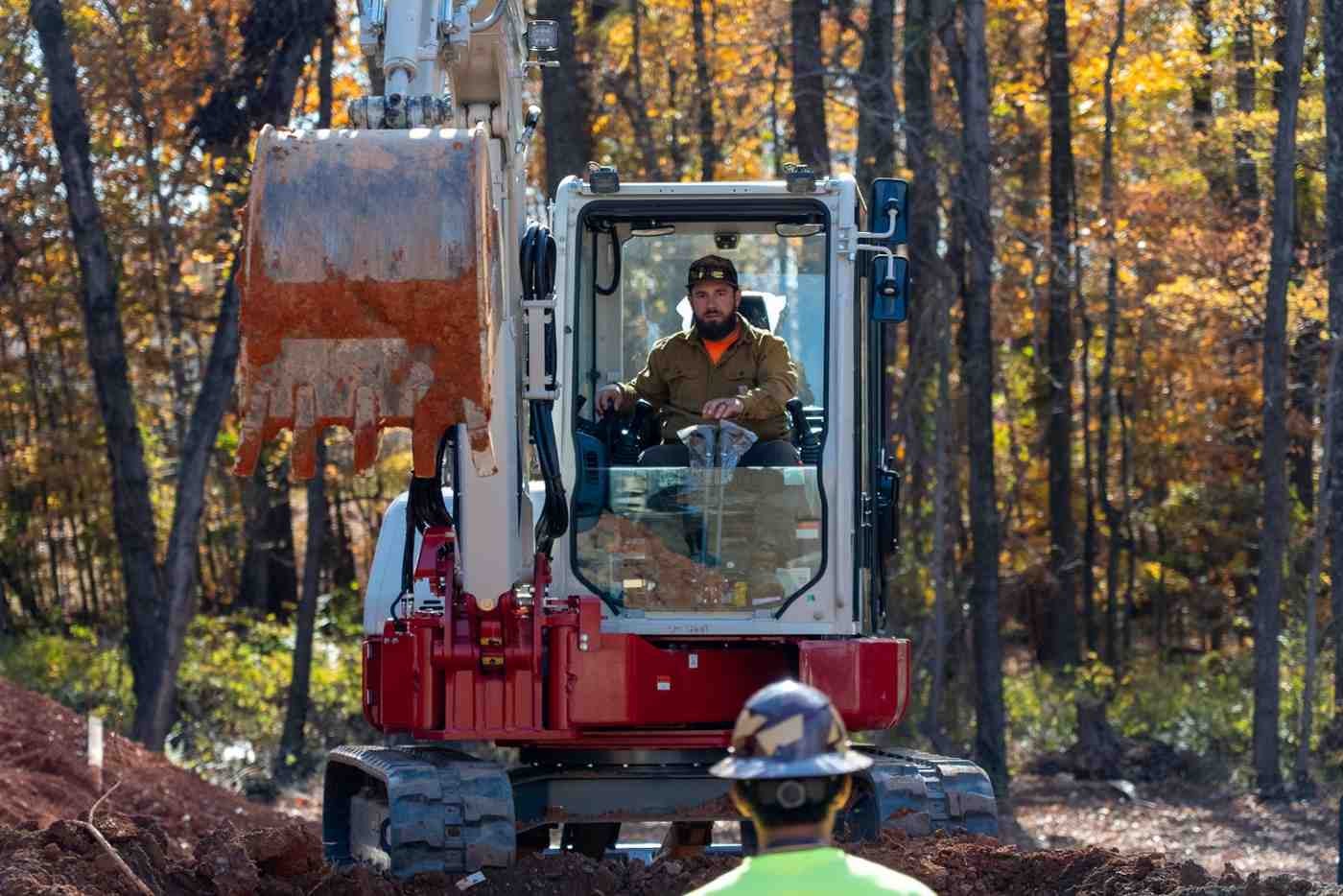 A man in a brown shirt and cap operates an excavator, its bucket full of dirt, with another man in a hard hat watching from the foreground.