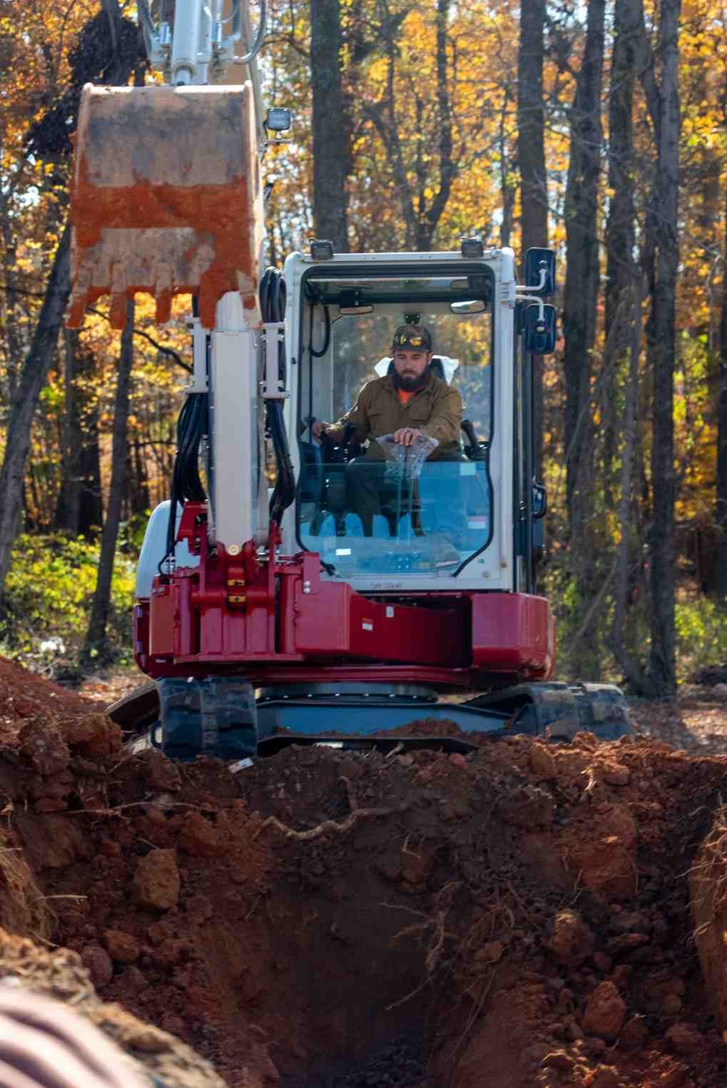 A bearded man in a baseball cap operates a red and white mini excavator digging a trench in a forest.