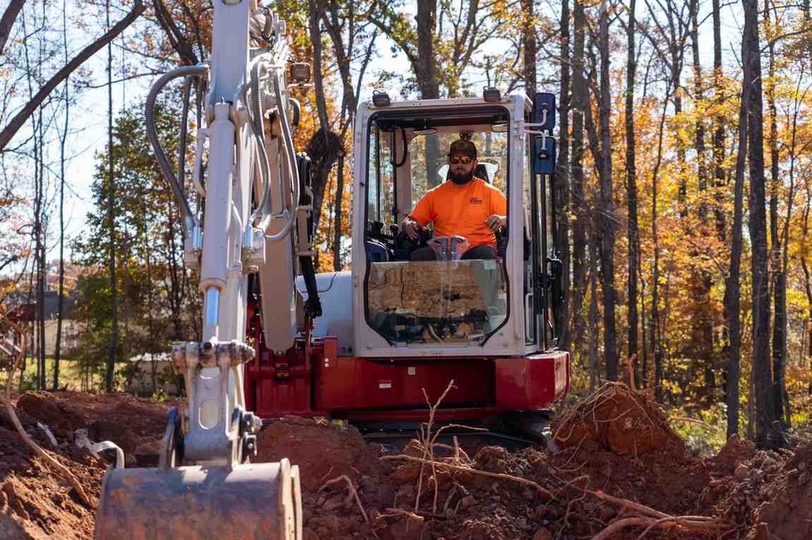 A man in sunglasses and an orange shirt operating an excavator on a sunny autumn day in a wooded area.