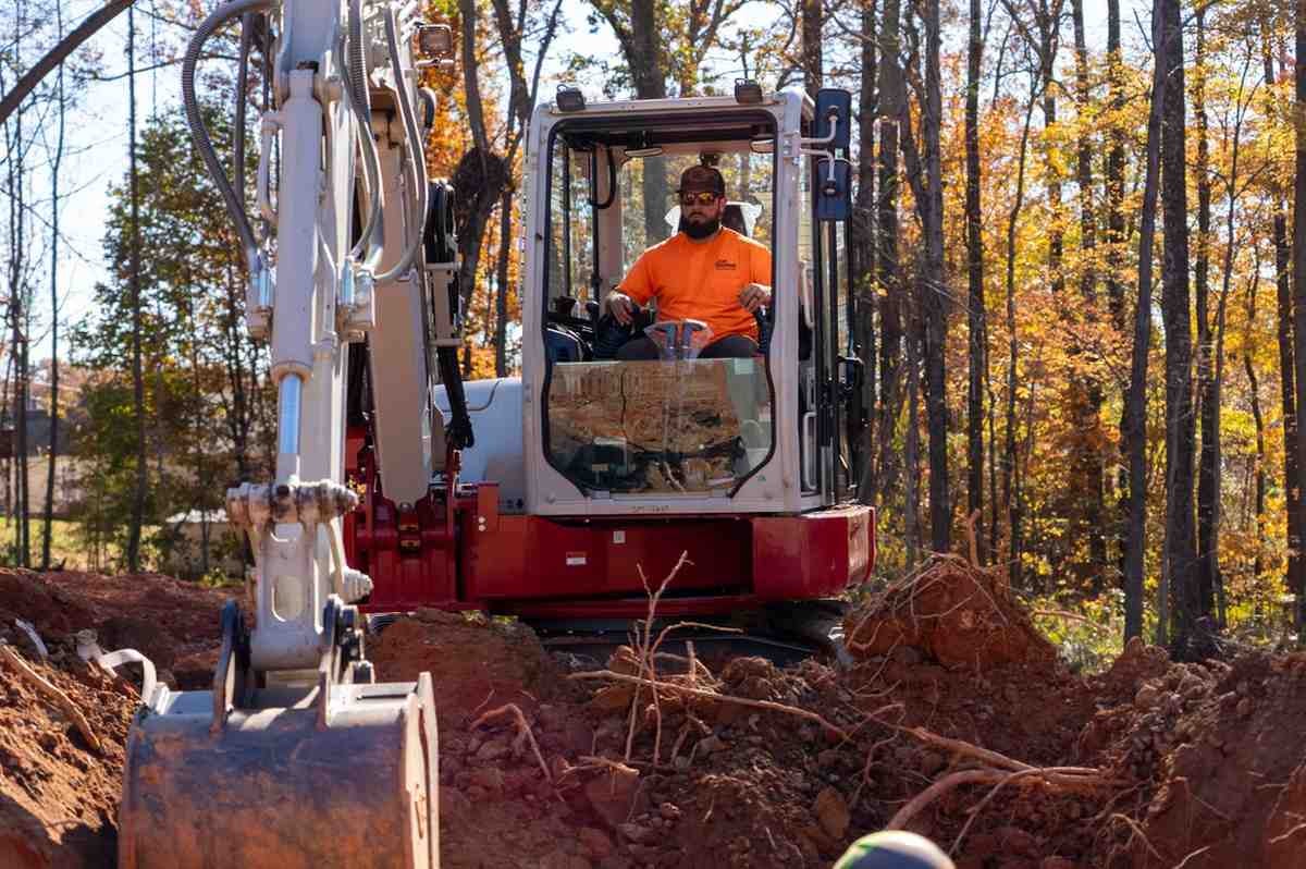A man in an orange shirt and sunglasses operates a white and red excavator digging a ditch in a forest.