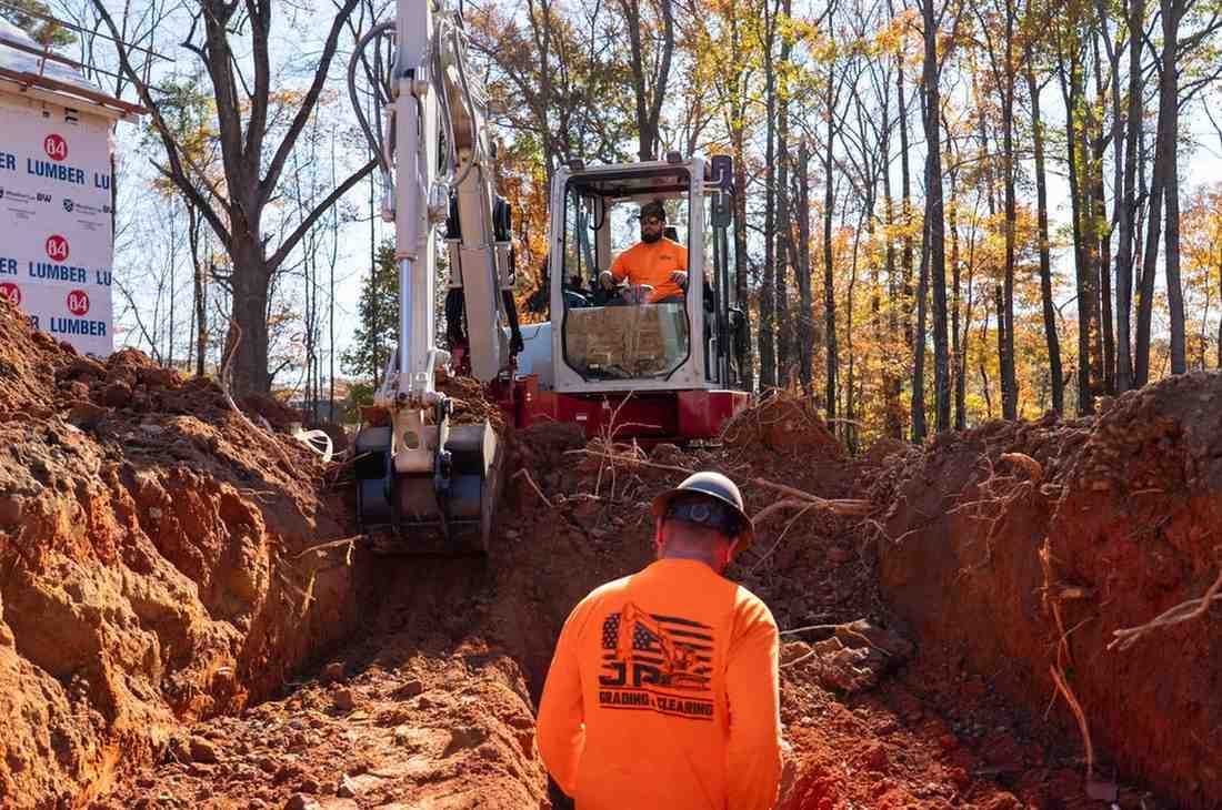 Two construction workers, one in an excavator and another watching from a trench, prepare a new home site.