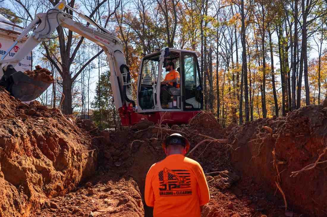 Two construction workers: one operates an excavator in a deep trench, while another views the trench from above.