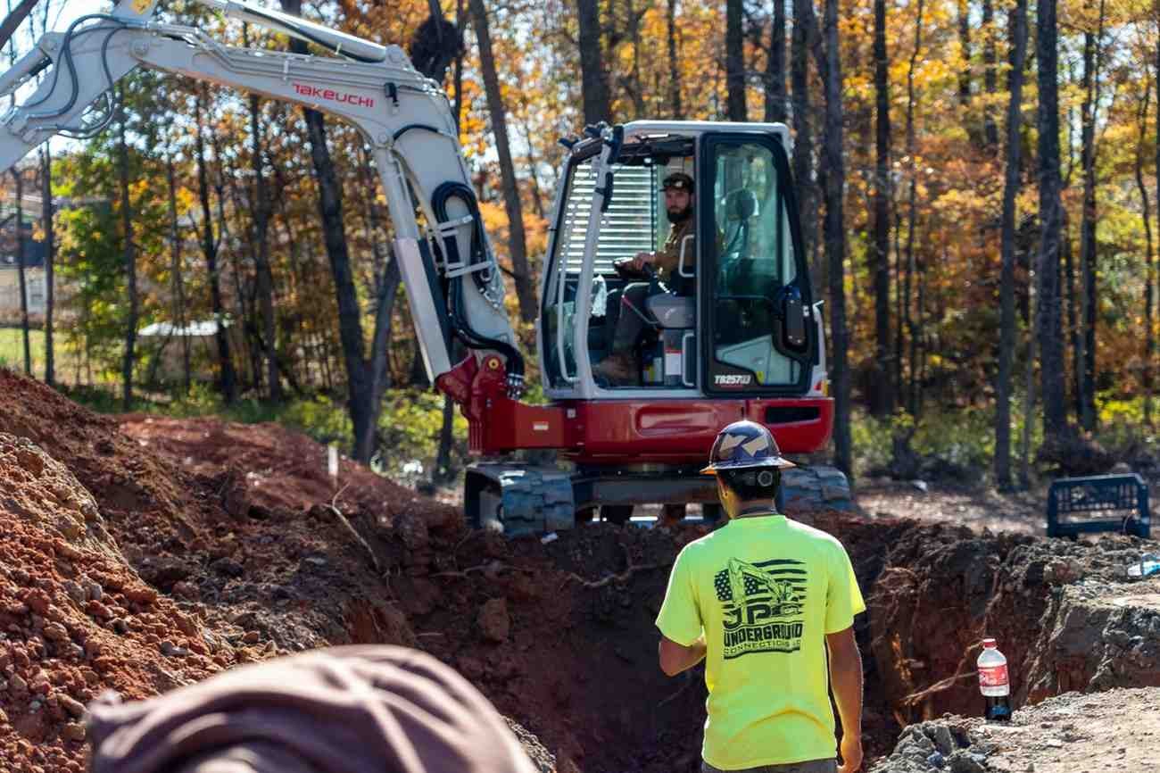 Two construction workers operating an excavator and working in a dirt trench, surrounded by trees.