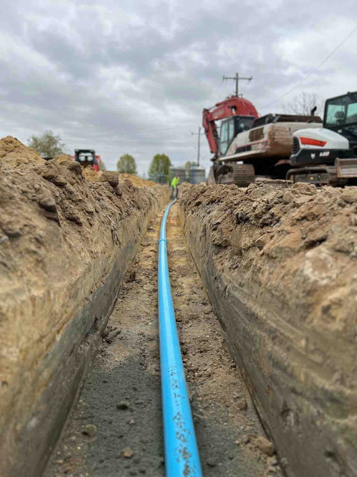 A long, narrow trench in dirt with a light blue pipe running down its center. Heavy equipment in background.