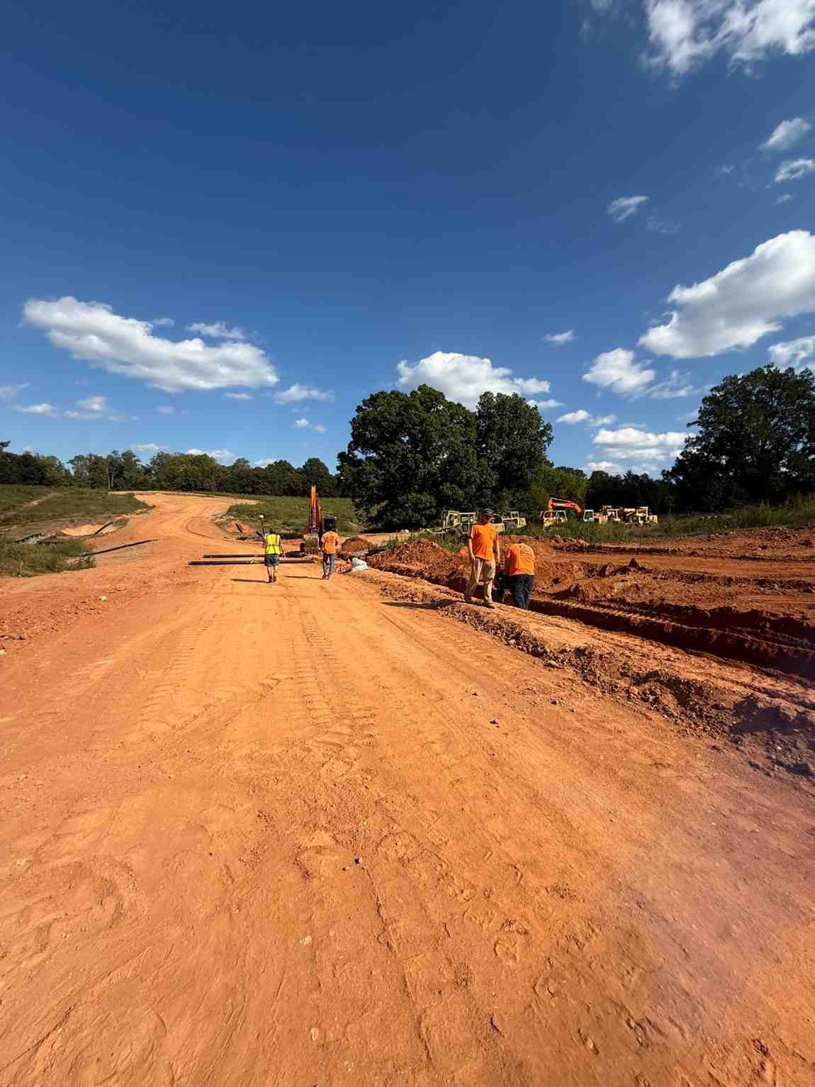 Construction workers and heavy machinery on a red dirt road project under a bright blue sky with white clouds.