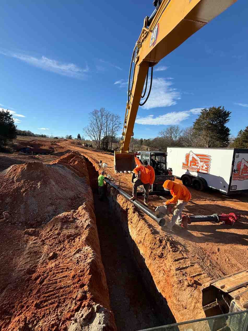 Construction workers in orange shirts laying a large pipe into a deep trench next to an excavator and trailer.