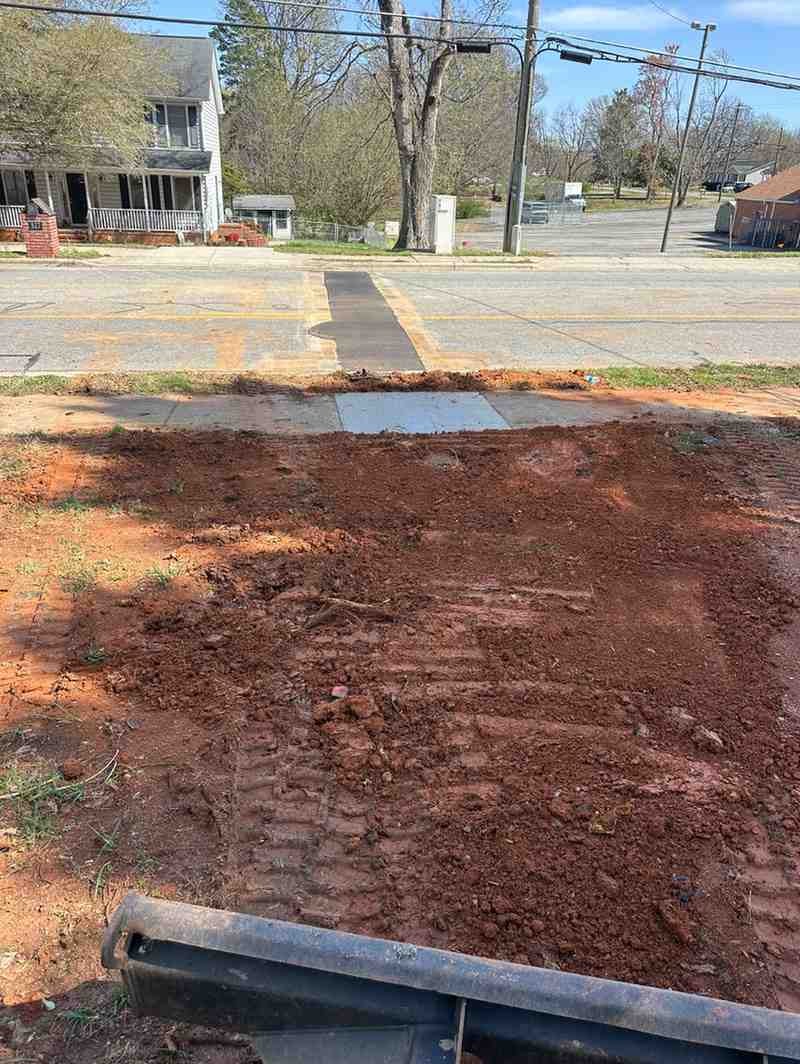 A dirt and mud construction site in the foreground, with a paved road and white house in the background.