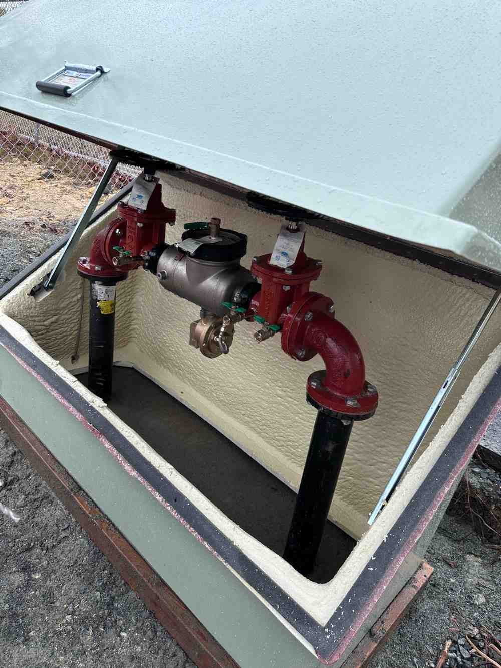 Open outdoor utility box revealing insulated walls, red valves, and black pipes on a gravel ground.