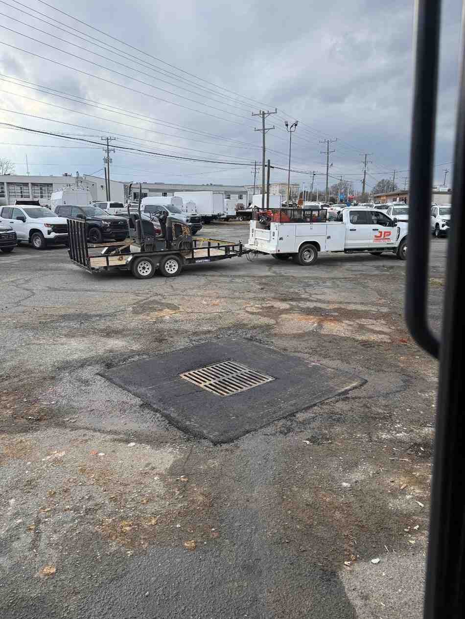 Outdoor parking lot with a newly paved square around a storm drain, surrounded by work trucks and trailers.