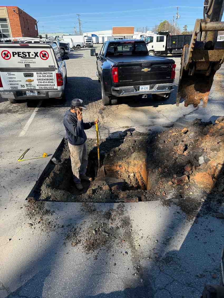 A man on his phone stands in a deep pit in an asphalt parking lot, next to a black pickup truck and an excavator.