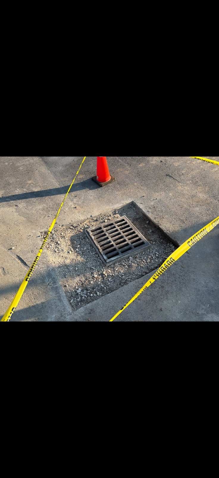 A storm drain surrounded by loose gravel, marked by yellow caution tape and an orange traffic cone.