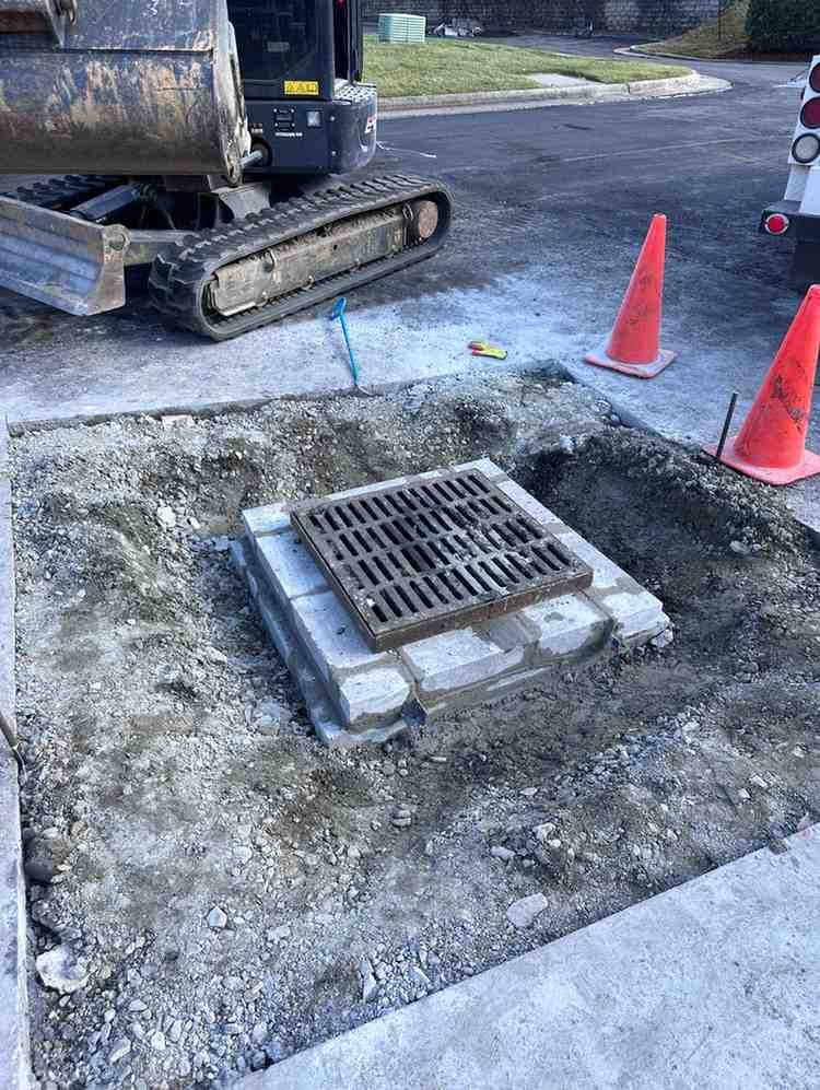 A new street drain with a metal grate, surrounded by gravel and concrete, next to construction equipment.