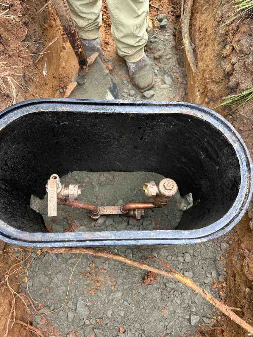A person in boots and tan pants stands over an open pipe access box in a ditch, exposing the copper pipes.
