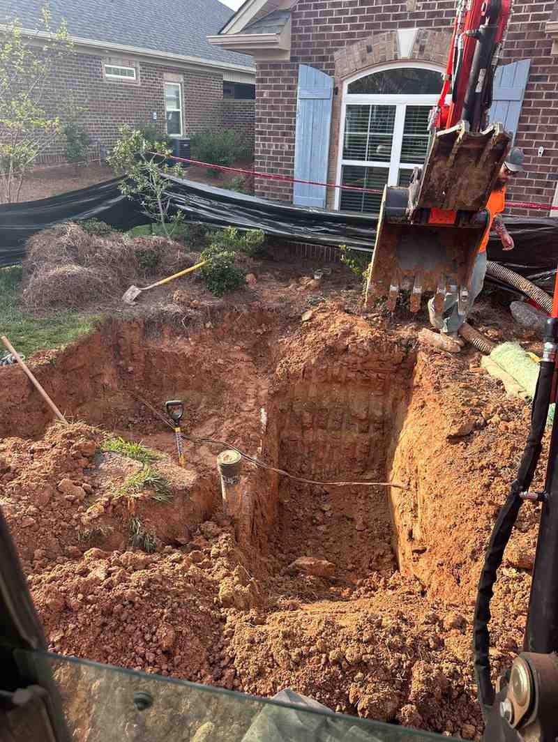 Excavator bucket over a large utility trench with exposed pipes and wires next to a brick house.