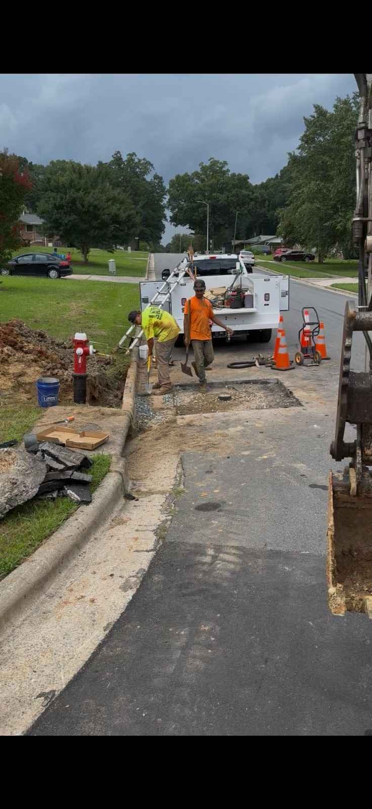 Two construction workers, one in orange and one in yellow, repair a curb next to a red fire hydrant and truck.