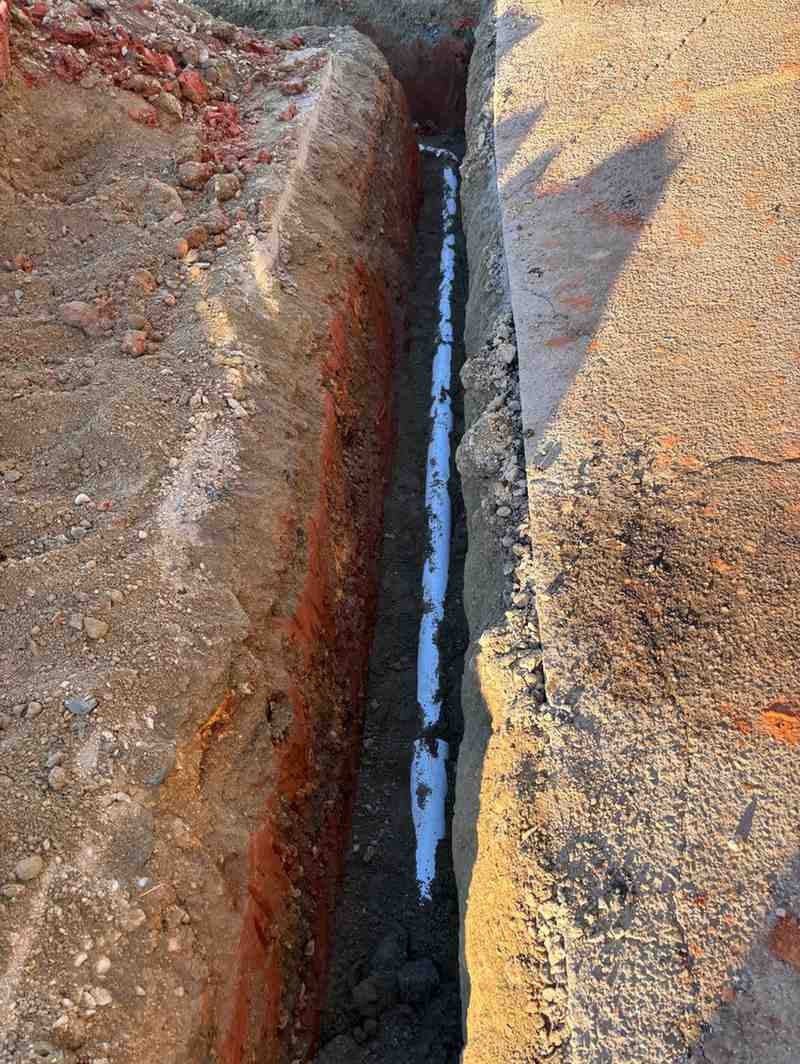 A narrow trench in the earth, revealing a white pipe running along its bottom.