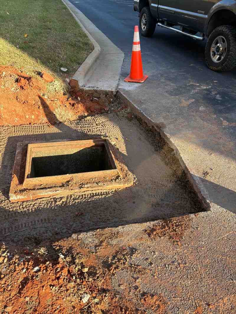 Open utility access hatch in a dirt trench next to a concrete curb with an orange cone and parked SUV.