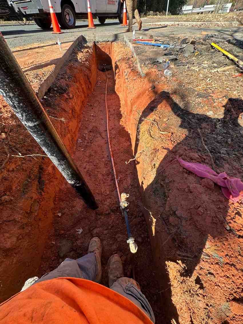 Worker standing in a deep trench with exposed pipes and utilities, construction vehicle in background.