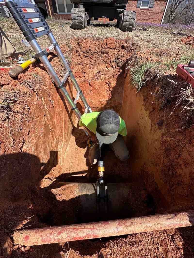 A person in a trench working on a pipe, with a ladder leaning against the side and an excavator in the background.